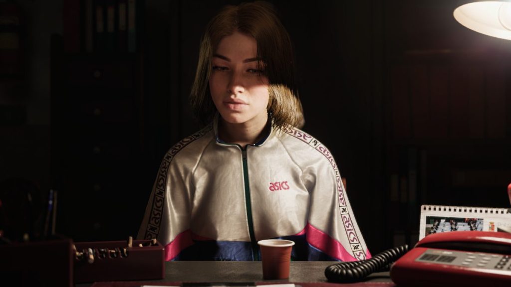 Femme assise devant un bureau, portant une veste ASICS, avec une expression pensive. Un petit gobelet est posé devant elle, à côté d'un téléphone rouge et d'un carnet.
