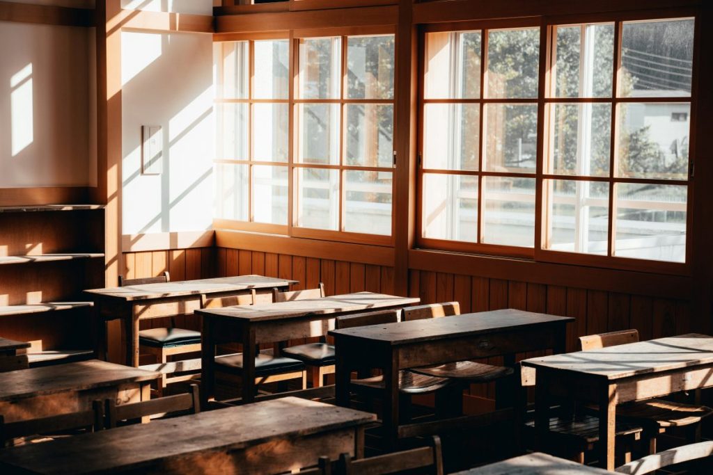 Intérieur d'une salle de classe avec des bureaux en bois et de grandes fenêtres laissant entrer la lumière naturelle.