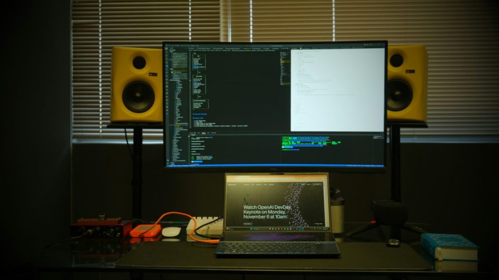 Bureau avec un ordinateur portable et un écran grandiose, affichant du code, accompagné de haut-parleurs jaunes.