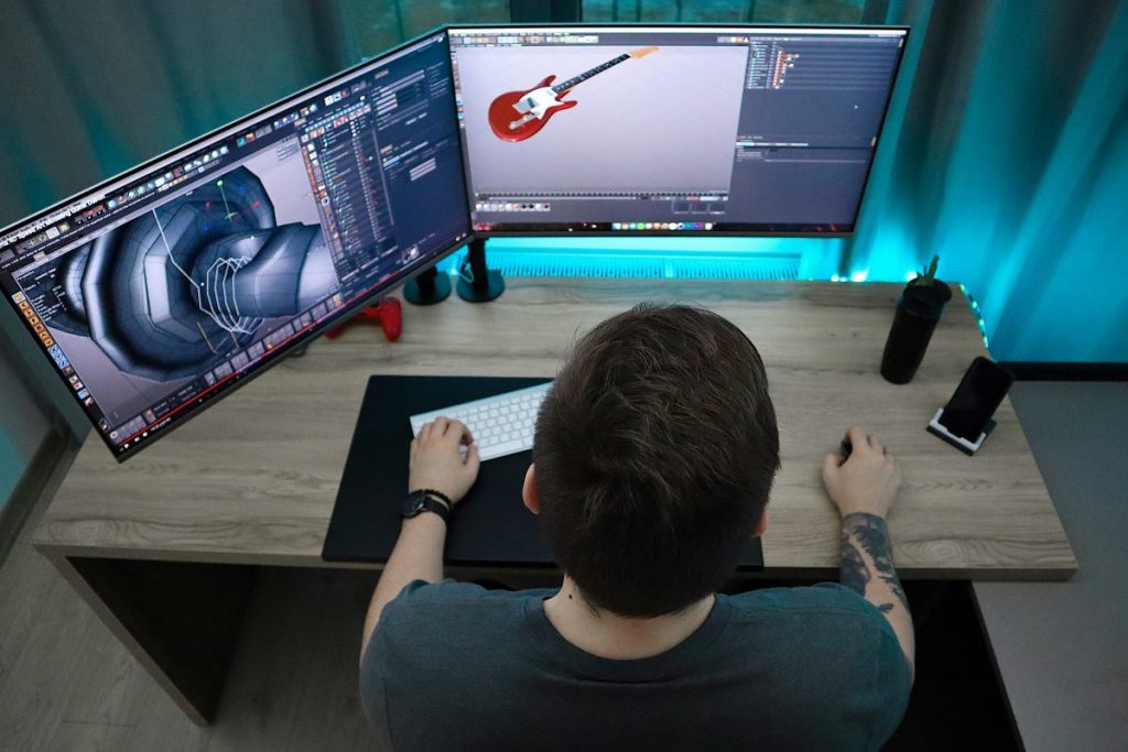 Un homme assis devant un bureau avec deux écrans d'ordinateur. L'écran de gauche affiche un logiciel de modélisation 3D, tandis que l'écran de droite montre une guitare en 3D. Le bureau est en bois, avec un téléphone portable et une tasse à côté.