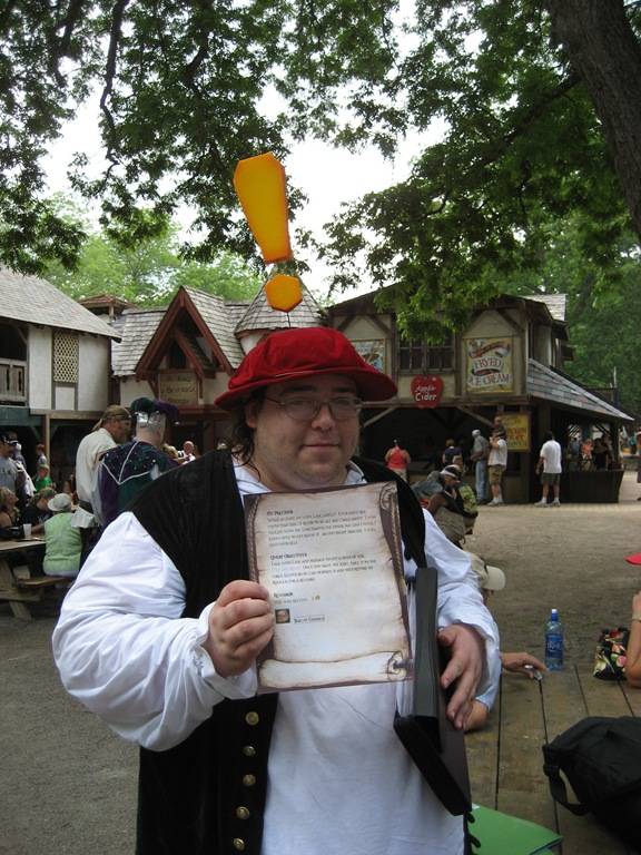 Un homme portant un chapeau rouge avec un panneau d'exclamation jaune, tenant un document dans un environnement de festival médiéval.