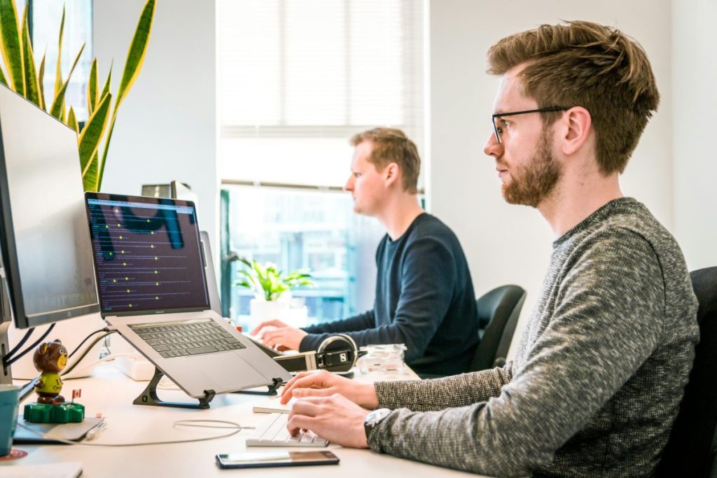 Deux hommes travaillant sur des ordinateurs portables dans un bureau lumineux, avec des plantes en arrière-plan.