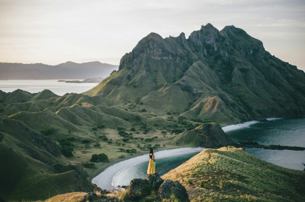 Une personne en robe jaune se tient sur une colline surplombant un paysage montagneux, avec une baie en arrière-plan au coucher du soleil.