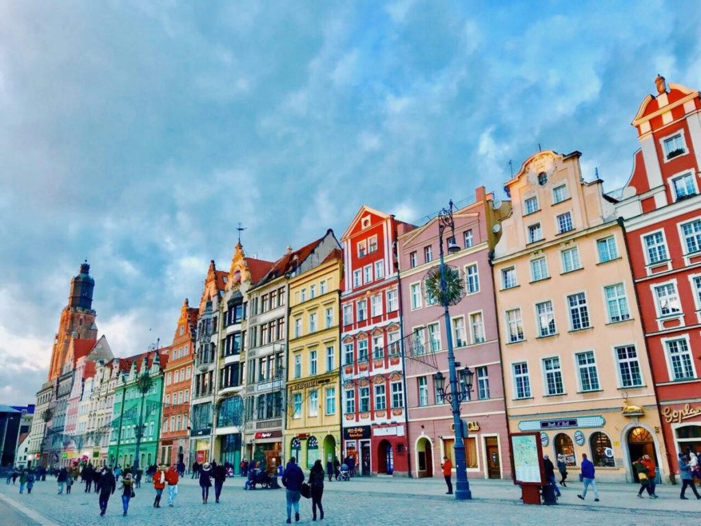 Vue colorée des bâtiments historiques sur une place, avec des personnes marchant sous un ciel nuageux.