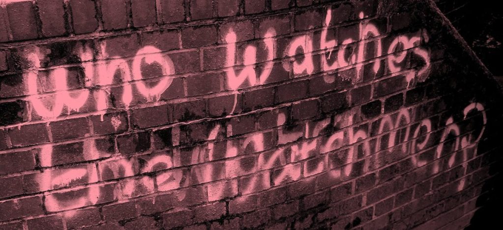 Graffiti sur un mur en briques, avec l'inscription 'Who watches the Watchmen?' en rose.