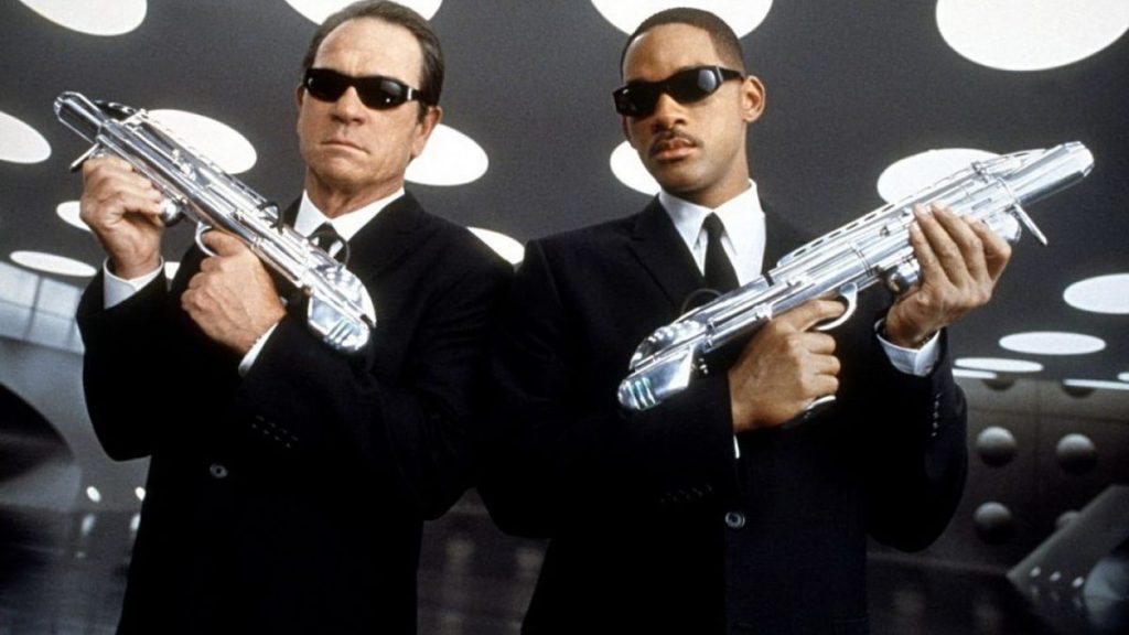 Deux agents en costume noir avec des lunettes de soleil, tenant des armes futuristes dans un environnement intérieur moderne.