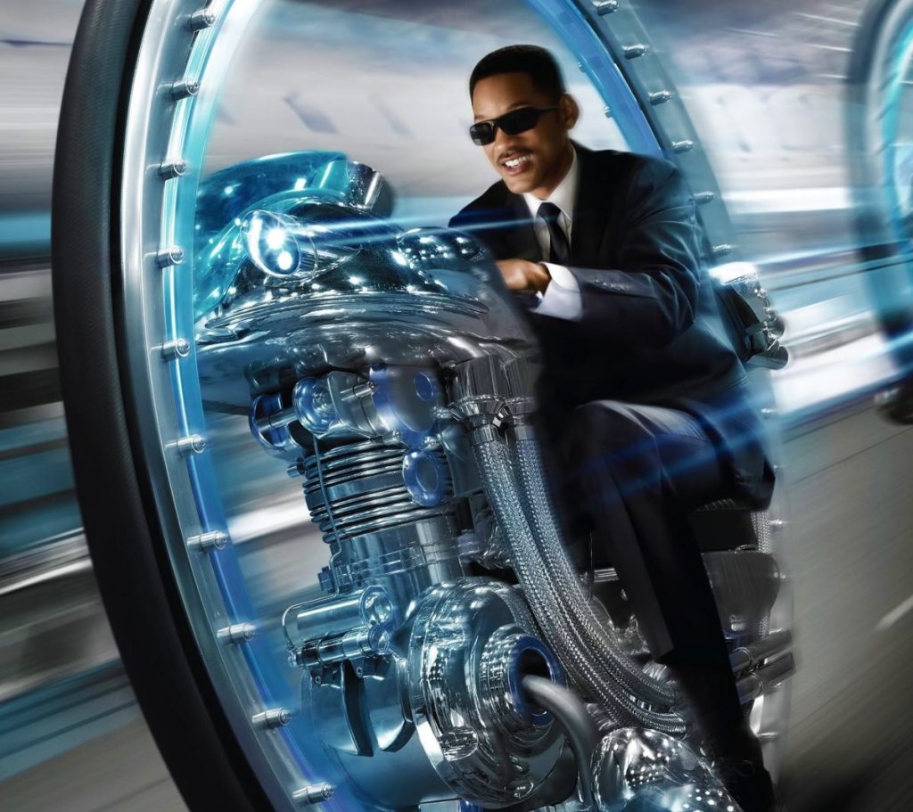 Un homme en costume noir, portant des lunettes de soleil, conduit une moto futuriste brillante dans un environnement dynamique.