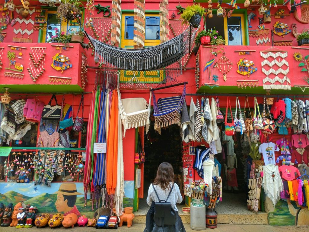 Une femme se tient devant une boutique colorée avec des vêtements et des artisanats, dont des hamacs et des pots en terre cuite, sur un mur peint de motifs vibrants.