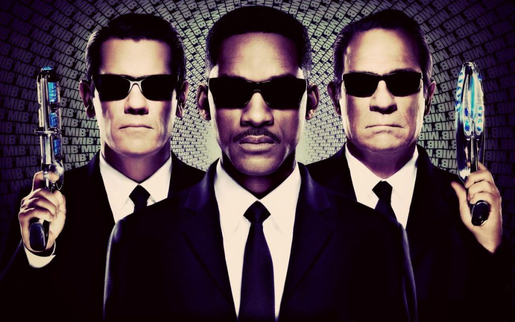 Trois hommes en costume noir avec des lunettes de soleil, tenant des armes futuristes, sur fond texturé avec des mots 'MIB'.