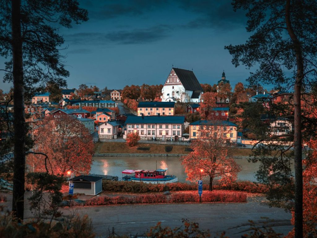 Vue panoramique d'une petite ville au bord de l'eau, avec des maisons colorées et une église distinctive, entourée d'arbres en automne.