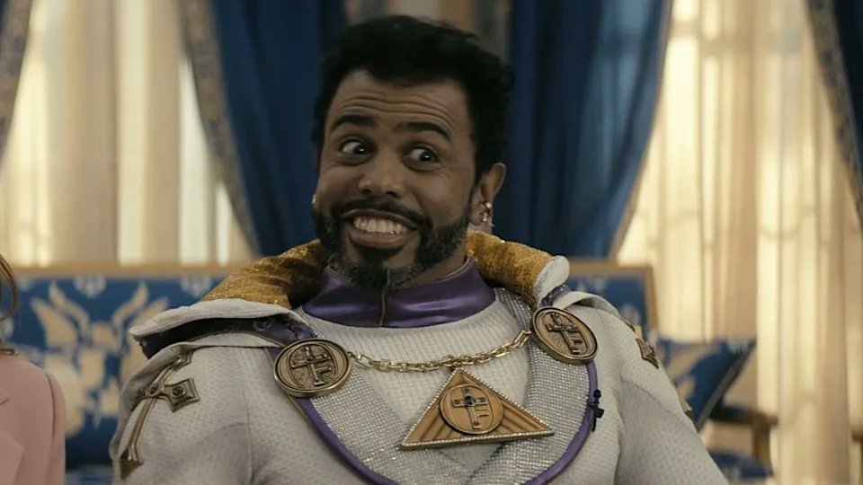 Un homme souriant portant un costume de super-héros avec des éléments dorés, assis dans un décor intérieur élégant.