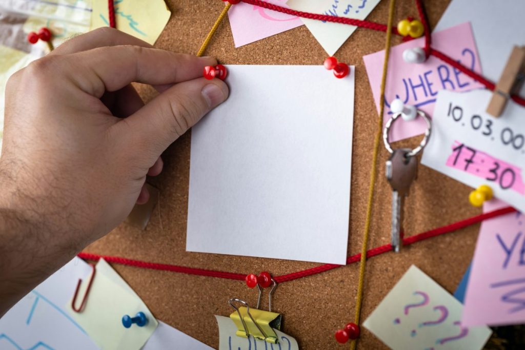 Une main tenant une note blanche qui est accrochée sur un panneau en liège, entourée de diverses notes colorées et de fils.