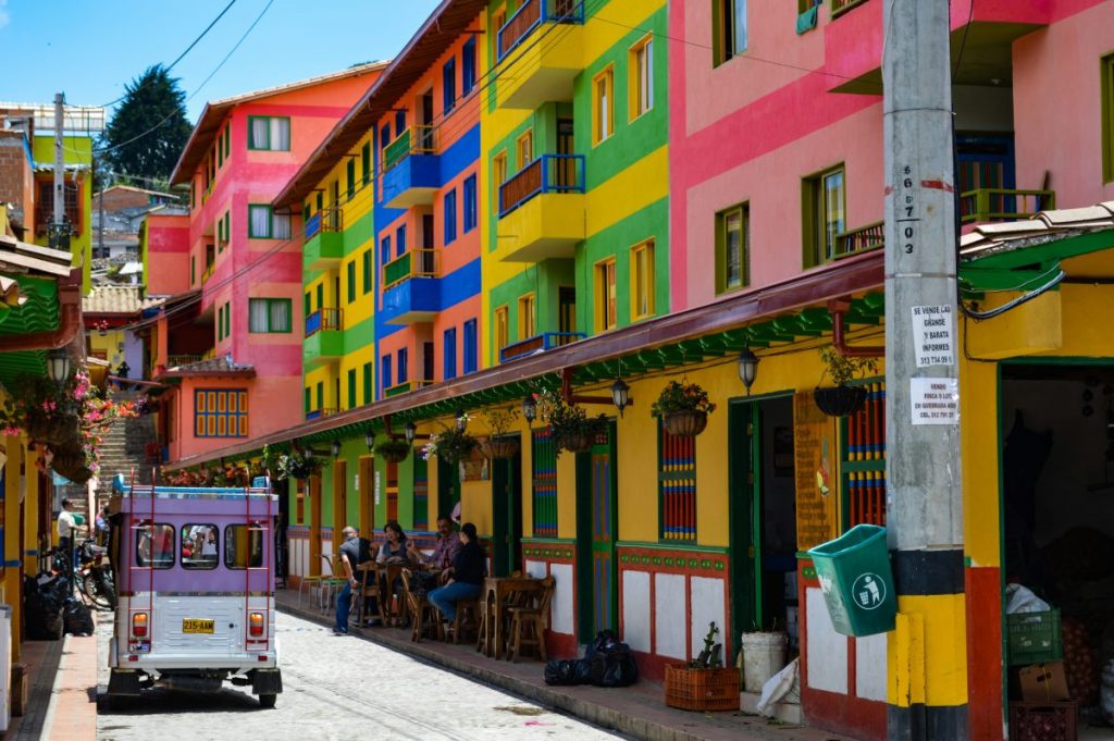 Une rue colorée avec des bâtiments aux teintes vives, incluant des murs rose, vert, jaune et bleu. Une voiturette blanche et mauve est garée, tandis que des personnes profitent des terrasses en bois.