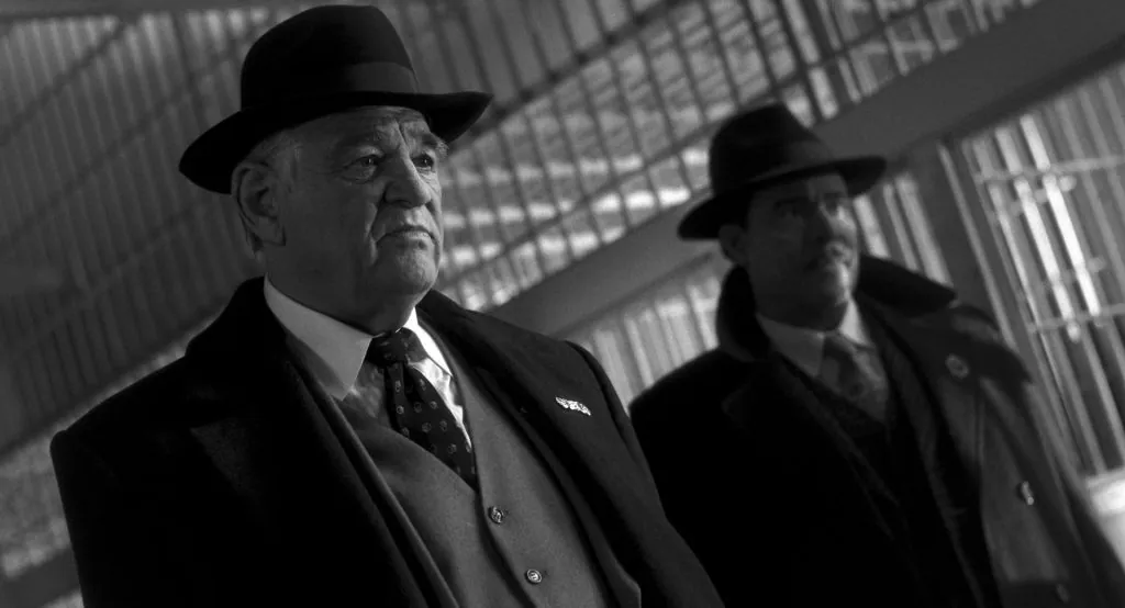 Deux hommes en costume avec des chapeaux, se tenant dans un cadre sombre et dramatique, en noir et blanc.